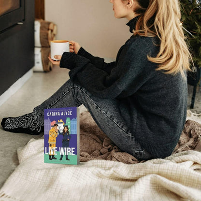 Woman sitting on a couch holding a mug with a book titled 'Live Wire' by Carina Alyce in front of her.