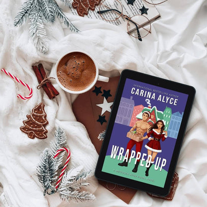 Tablet displaying a book cover titled 'Wrapped Up' by Carina Alyce, with a mug of hot chocolate and Christmas decorations on a white blanket.