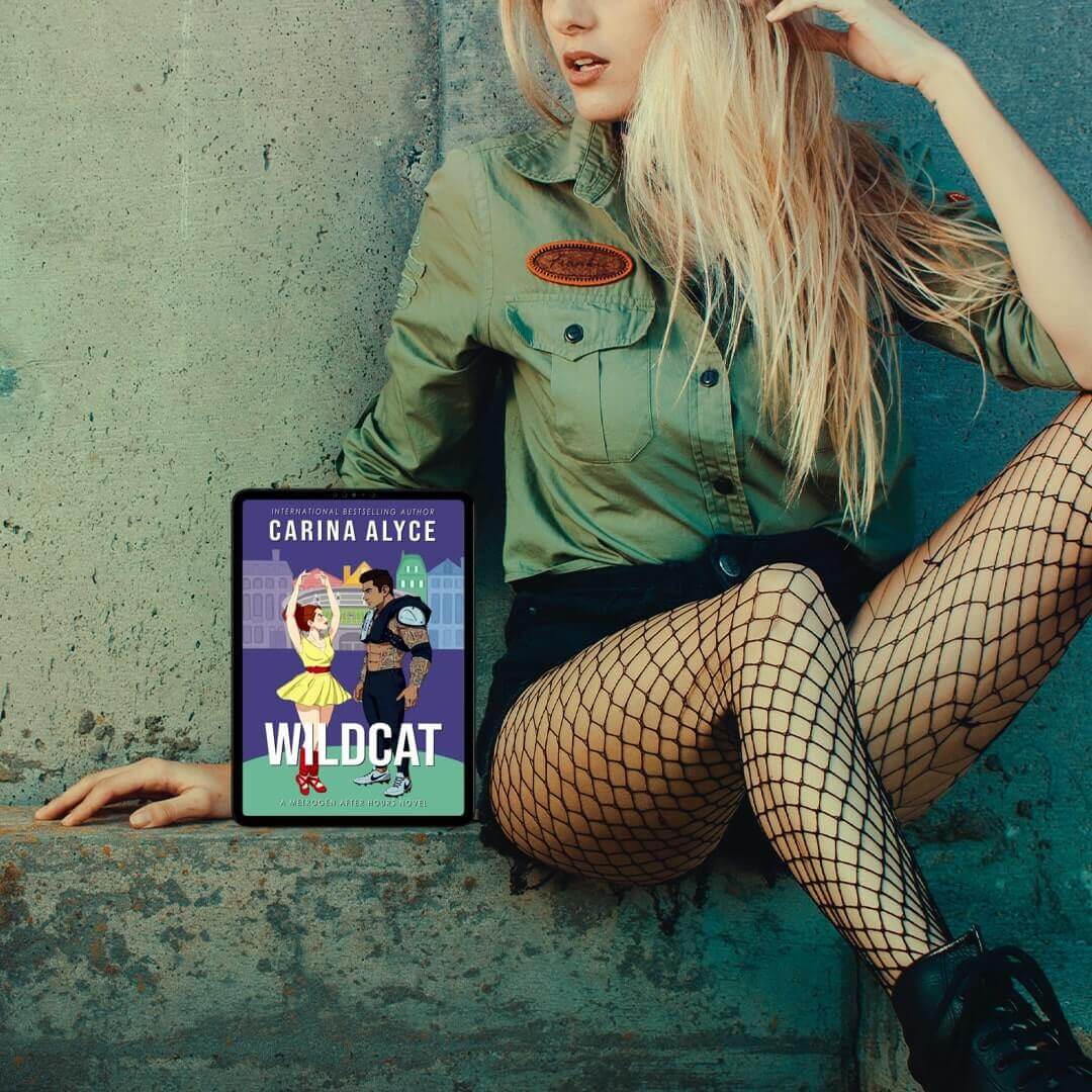 Woman in green shirt and fishnet stockings holding a book titled 'Wildcat' by Carina Alyce against a textured wall.