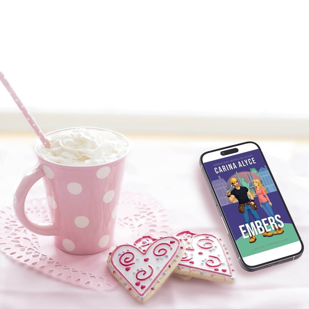 Pink polka dot mug with whipped cream, heart-shaped cookies, and a phone displaying 'Carina Alice Embers' on a white surface.