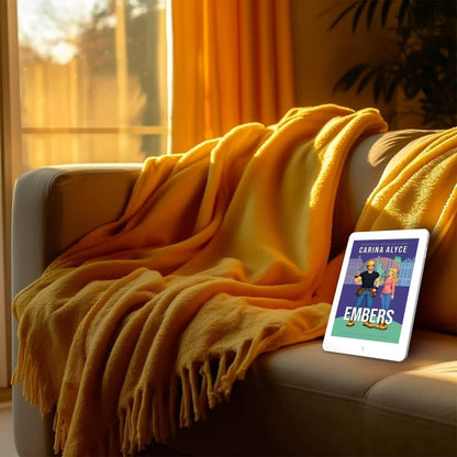 Yellow blanket draped over a couch with a tablet displaying a book cover titled 'Embers' by Carina Alyce.