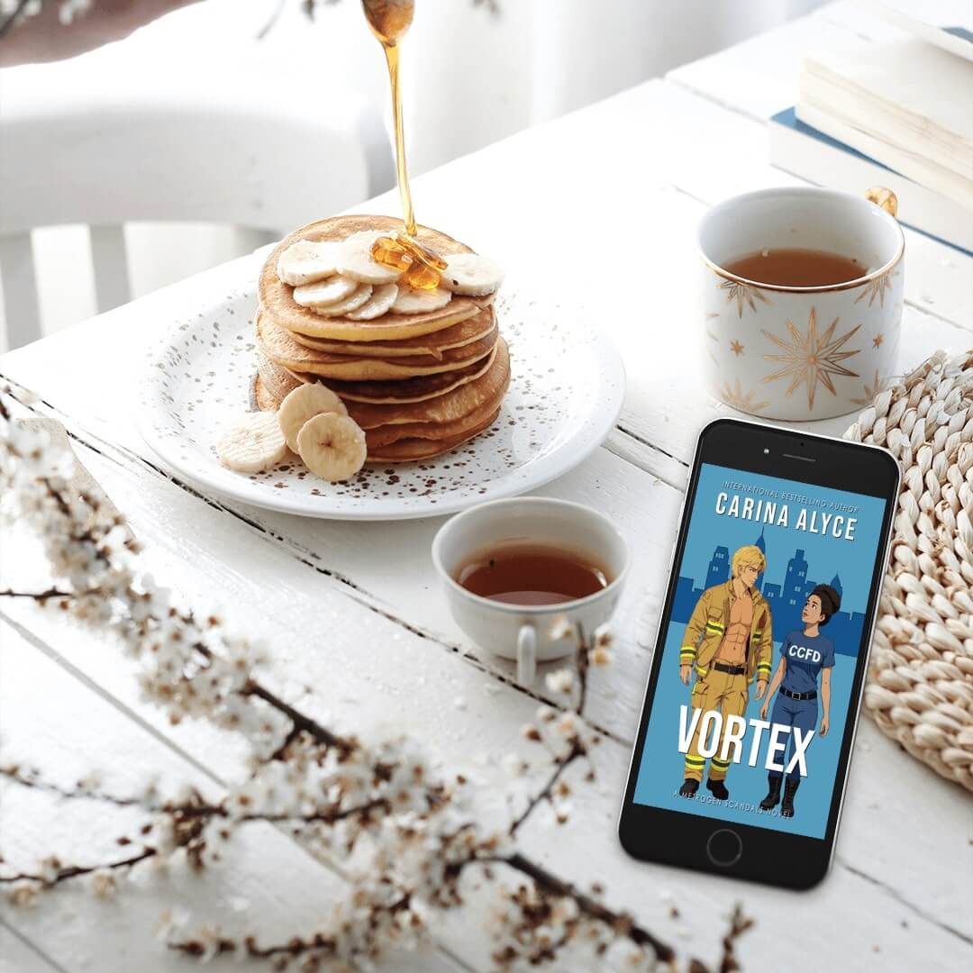 Stack of pancakes with syrup, bananas, and a cup of tea on a white table with a phone displaying 'Vortex' by Carina Alyce.